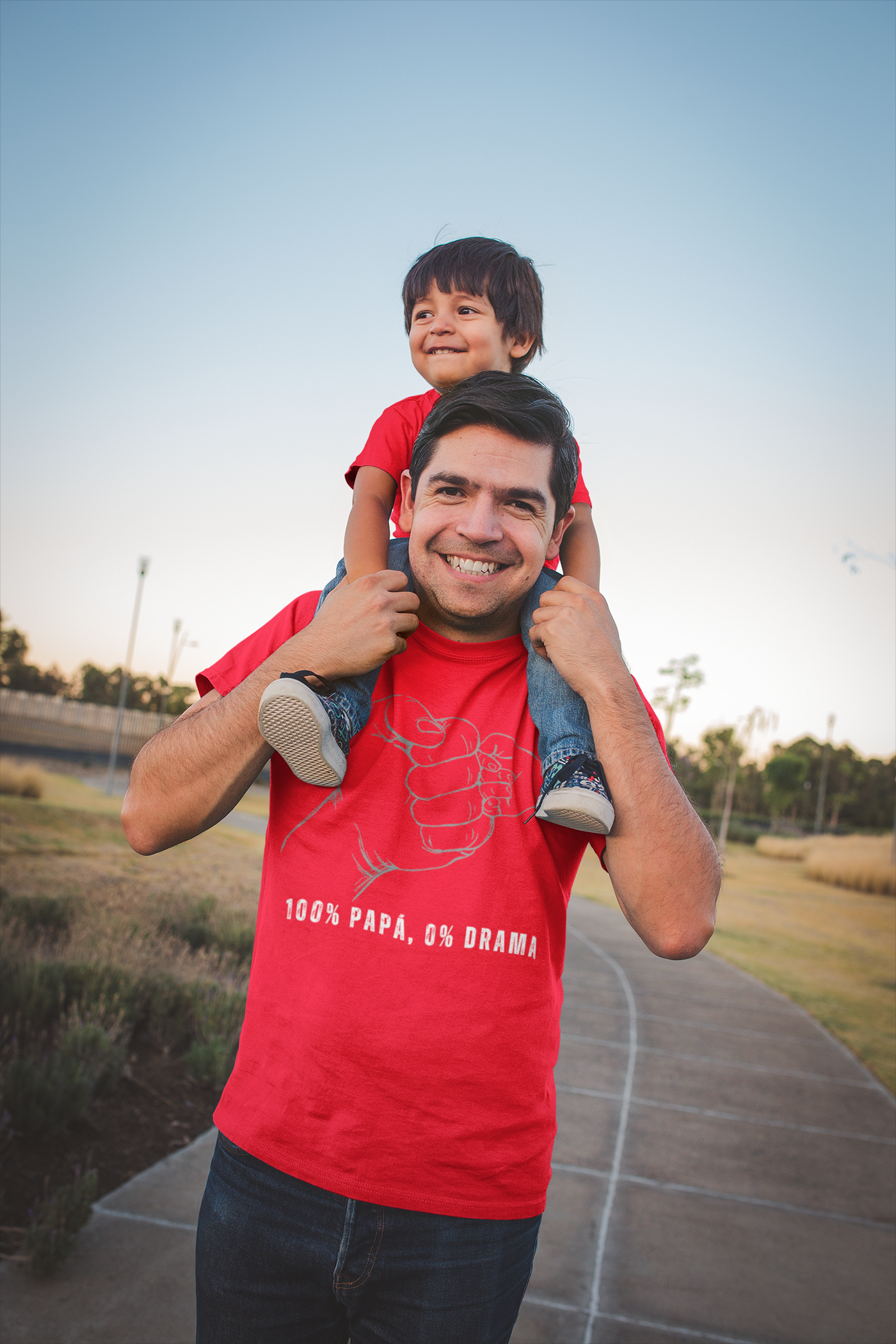 Man wearing "100 Percent Papá, 0 Percent Drama" shirt carrying a child on shoulders outdoors.